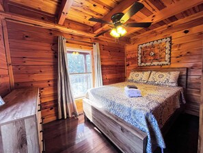 Ein Schlafzimmer einer Holzhütte mit einem Bett, einem Deckenventilator, einem Fenster mit Vorhängen und einem gerahmten Bild an der Wand.