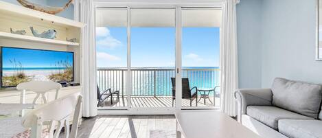 A spacious balcony with a view of the ocean, a white sofa, a dining table, and a TV on a shelf.