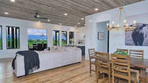 Ein moderner Wohnraum mit Sofa, Esstisch und Stühlen. Es gibt einen Deckenventilator, eingelassene Beleuchtung und einen Blick ins Freie durch große Fenster.