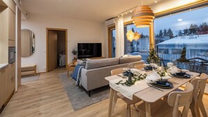 Moderni olohuone, jossa on sohva, kahdelle henkilölle katettu ruokapöytä ja taustalla televisio.