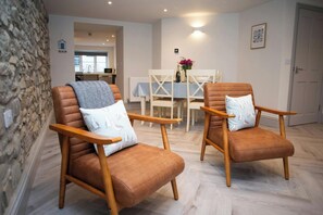 Two brown leather chairs with wooden armrests and cushions, a stone wall, a dining table with a blue tablecloth, and a kitchen in the background. - Bay View, New Quay - Two Bedroom Cottage, Sleeps 4 (New Quay)