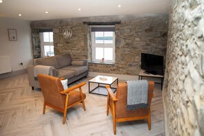 Un salon avec un mur en pierre, un canapé, deux fauteuils, une table basse et un téléviseur.