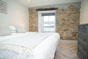 Une chambre avec un mur en pierre, un lit avec une literie blanche, une commode et une fenêtre donnant sur un beau paysage.