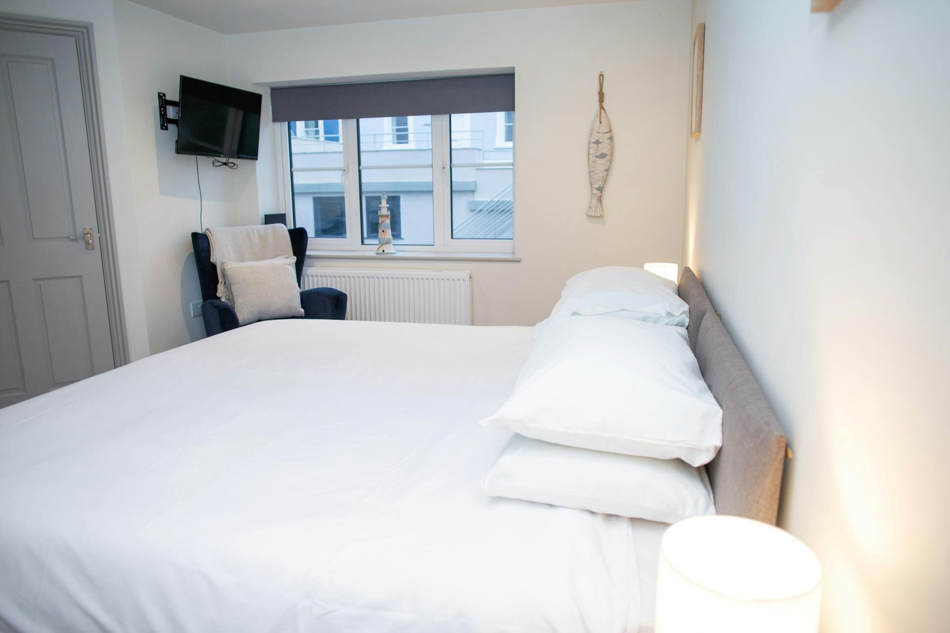 A bedroom with a bed, a chair, a television mounted on the wall, and a window with a view of buildings.