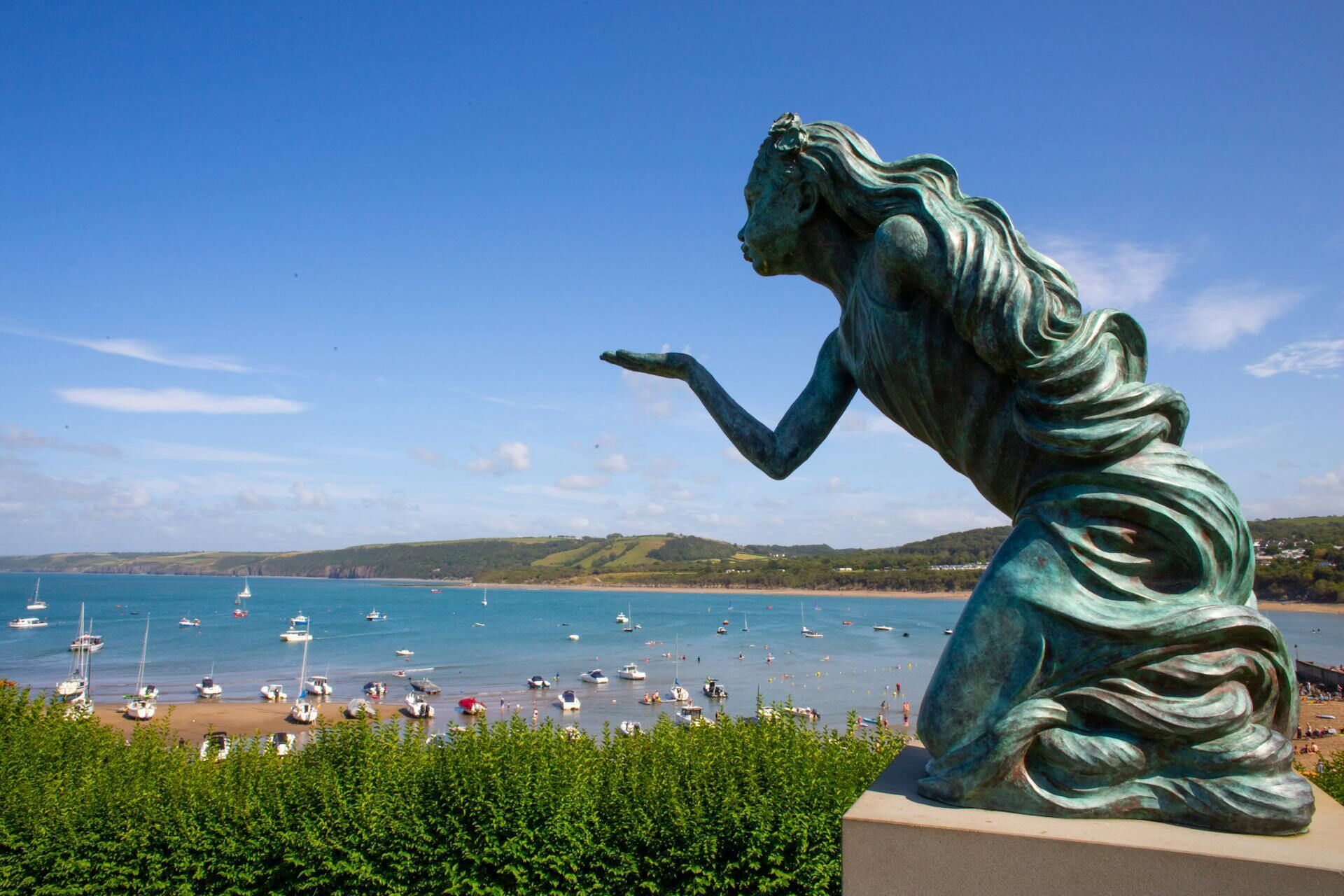 Une statue de bronze surplombant une zone côtière, avec des bateaux et une plage.