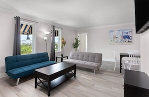 A modern living room with a blue sofa, a grey sofa, a wooden coffee table, and a dining area with white chairs and a small table. - Snowbird 2BR Condo On The Boardwalk + 100 Dollar Resort credit (Myrtle Beach)
