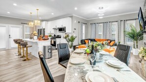 Une cuisine moderne avec des armoires blanches, un réfrigérateur en acier inoxydable et un four à micro-ondes. On y trouve un coin repas avec une table en marbre pour quatre personnes, des chaises noires et un canapé avec des coussins orange.