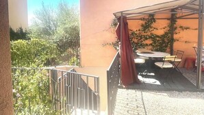 Een overdekt terras buiten met een tafel en stoelen, omringd door een metalen hekwerk en een tuin.
