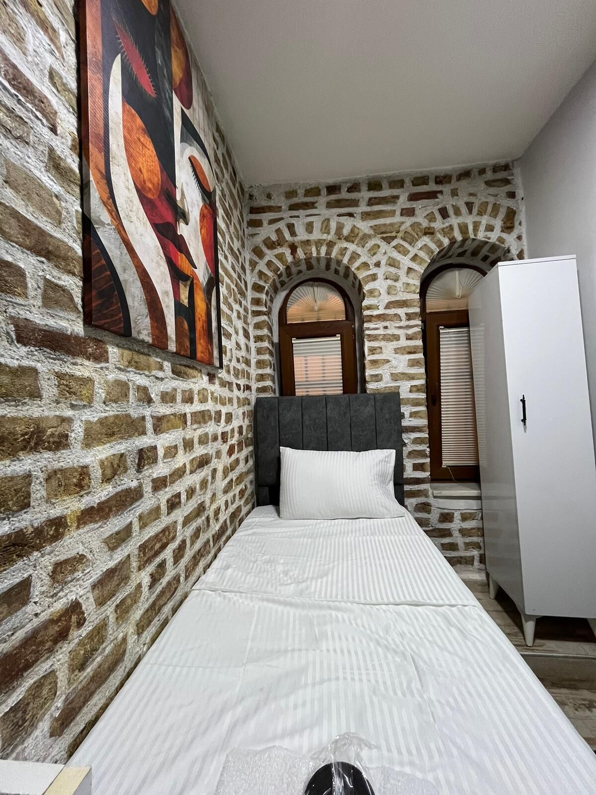 Een slaapkamer met een bakstenen muur, een bed met witte beddengoed, een grijs hoofdeinde en een witte garderobe.