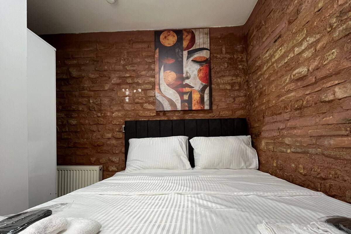A bedroom with a brick wall, a large abstract painting, a bed with white bedding, and a radiator.
