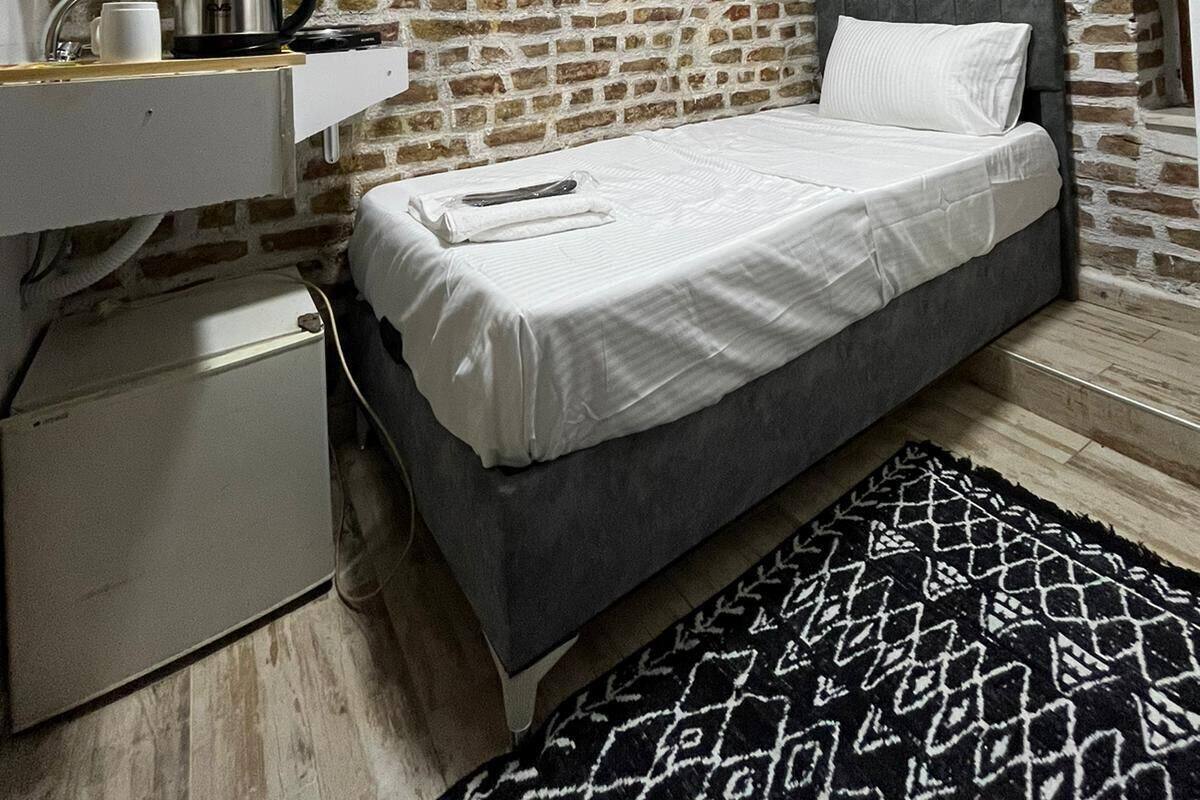 A compact bedroom with a brick wall, a bed with white linens, a small bedside table with a kettle, and a black and white patterned rug.