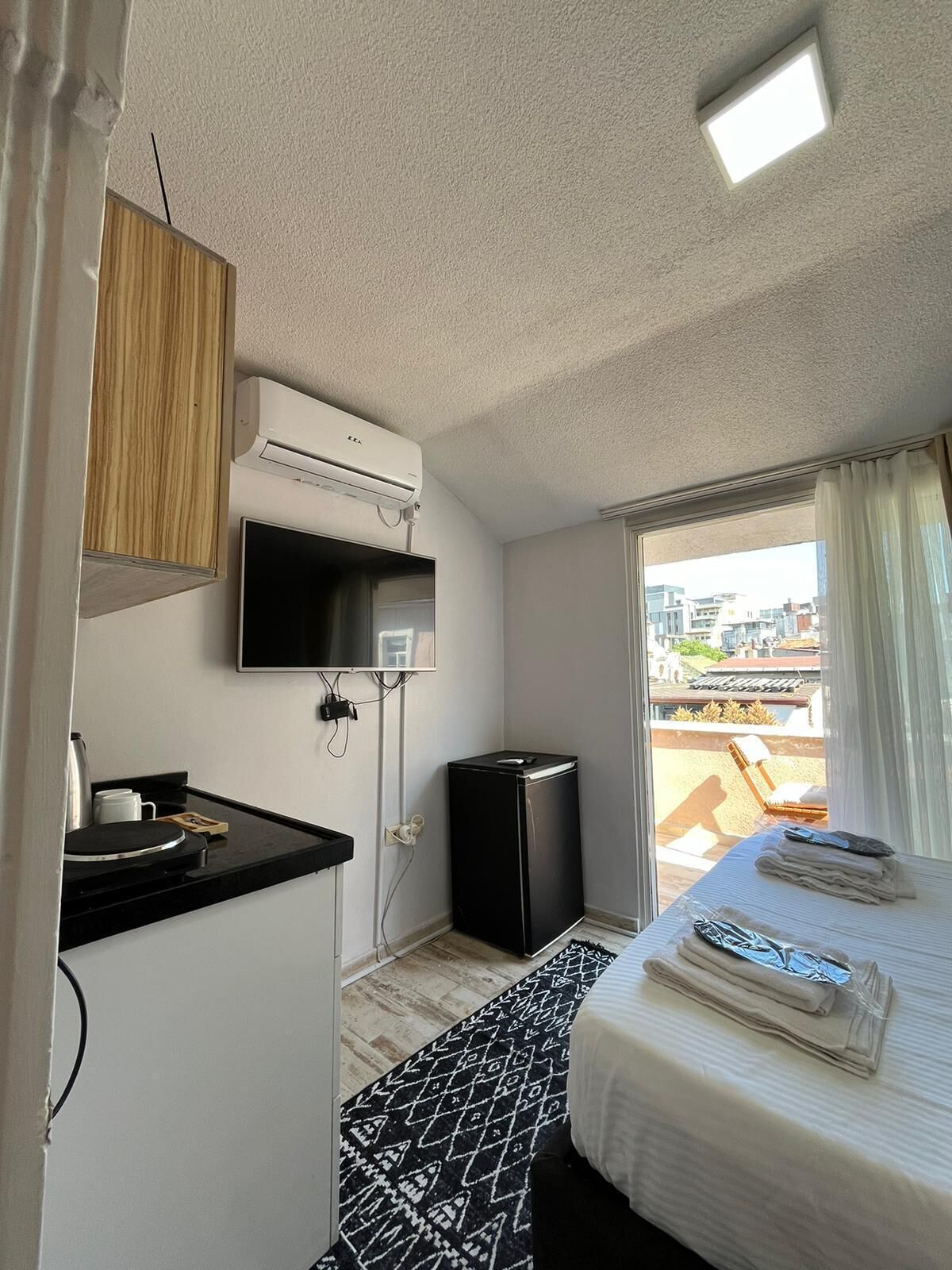 A compact hotel room with a bed, a small kitchenette, a TV, and a window with a view of buildings.