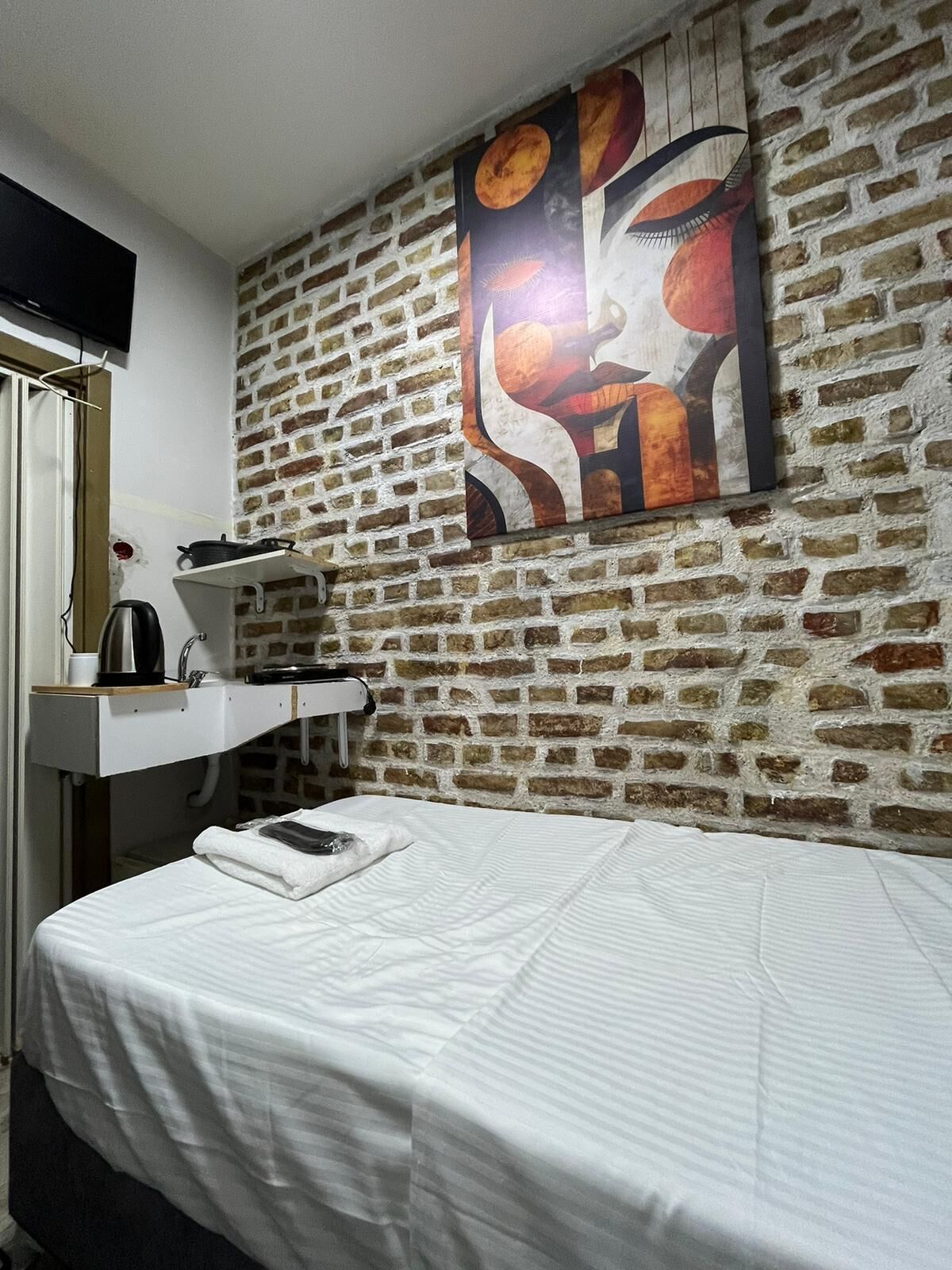 Habitación de hotel con pared de ladrillo, cama con ropa blanca, mesita de noche blanca con tetera y un cuadro sobre la cama.