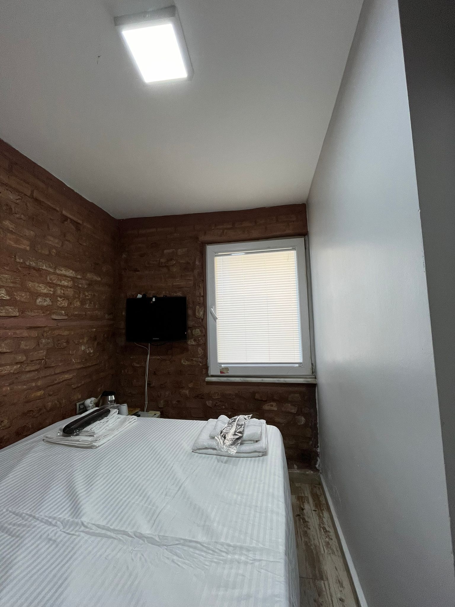 A small, compact room with a single bed, a flat-screen TV mounted on the brick wall, and a window with blinds.