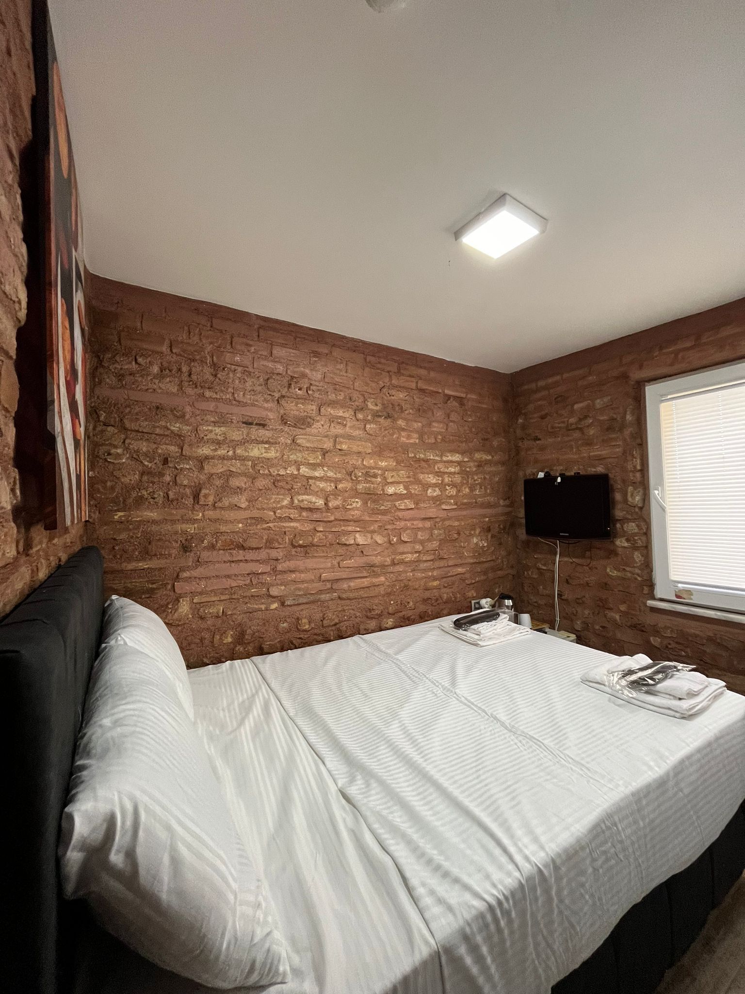 Een kamer met een bakstenen muur, een bed met wit beddengoed, een flatscreen televisie aan de muur en een raam met zonwering.