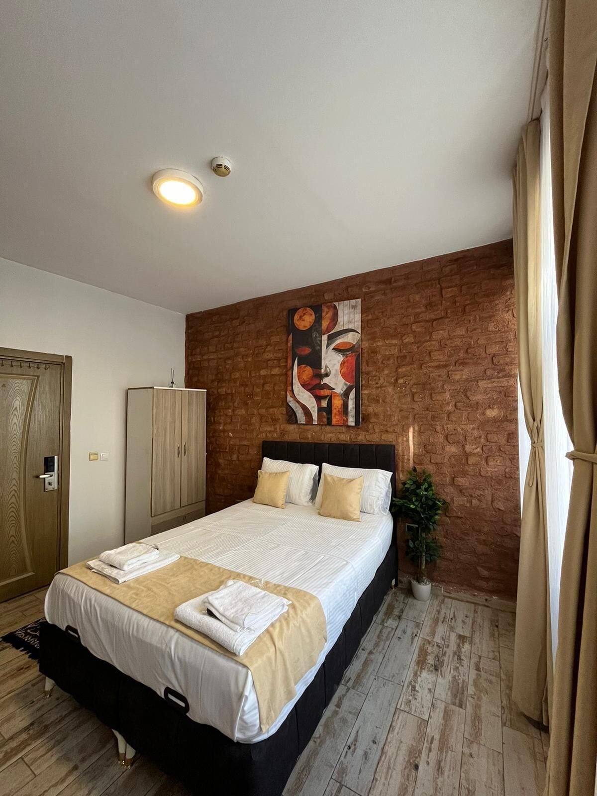Une chambre d’hôtel moderne avec un mur d’accent en briques, un grand lit avec une literie blanche, une armoire en bois et une plante en pot.