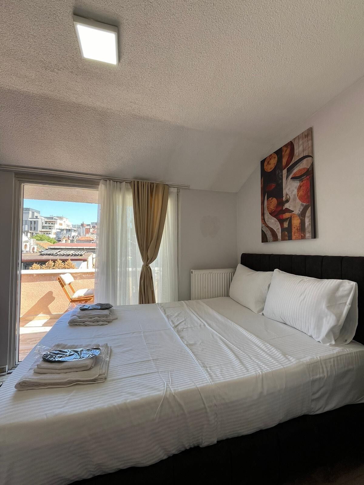 A hotel room with a bed, a radiator, a window with curtains, a view of buildings, and a ceiling light.
