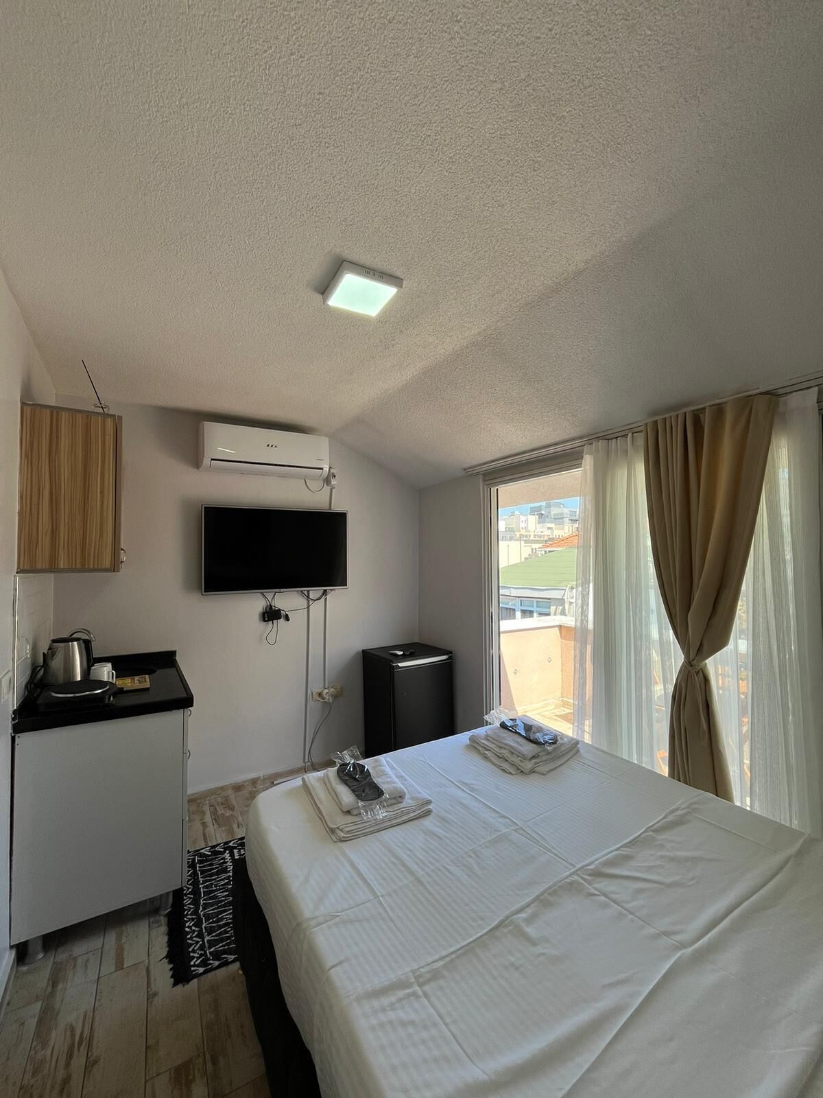 A hotel room with a bed, a TV, a desk with a kettle, a trash bin, and a window with curtains.
