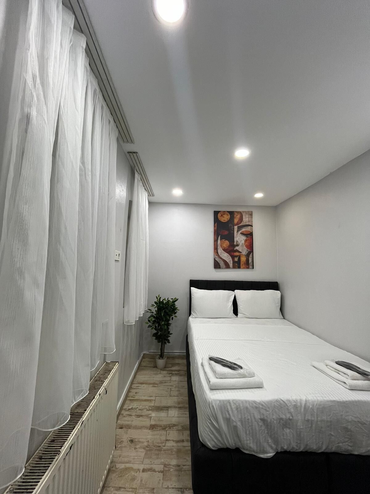 A modern hotel room with a large bed, white curtains, a potted plant, and a painting on the wall.