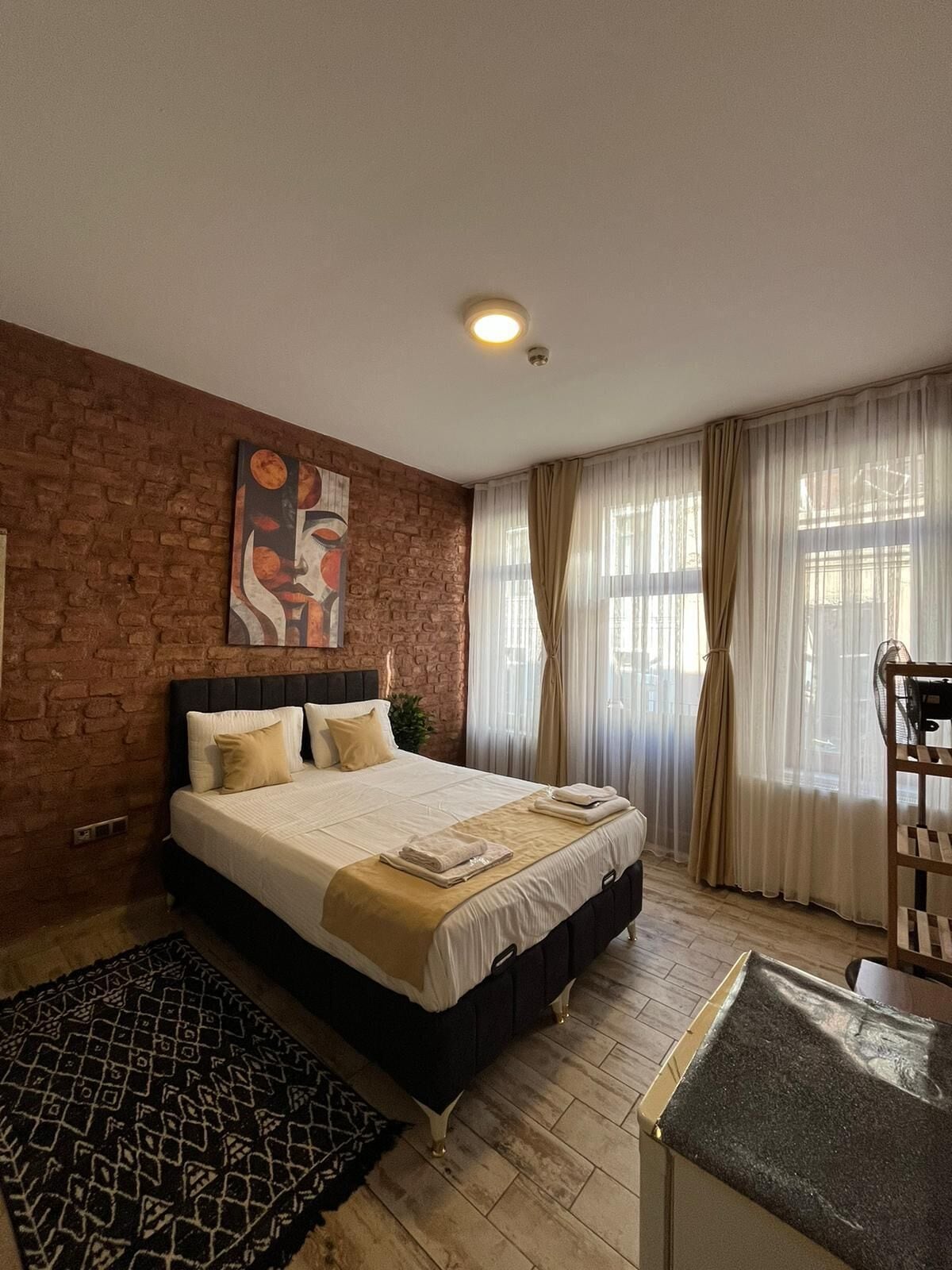Un dormitorio con pared de ladrillo, una cama con cabecera, una ventana con cortinas translúcidas y una pequeña mesa.