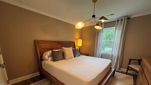 A bedroom with a wooden bed, a nightstand with a lamp, a window with curtains, and a view of greenery outside. - Quaint 2-bedroom apartment Historic downtown Florence (Florence)