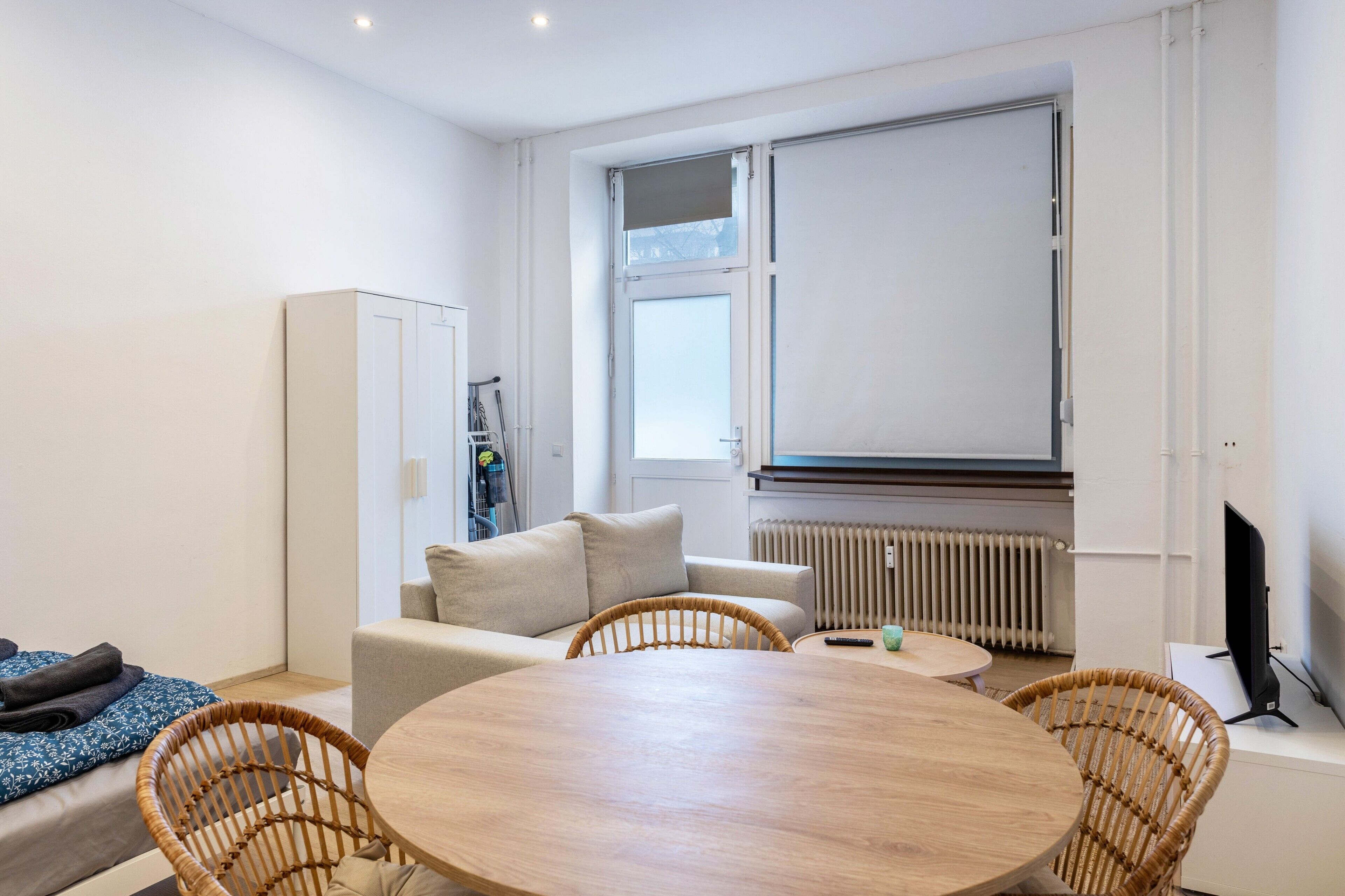A modern room with a round wooden table, wicker chairs, a sofa, a wardrobe, a radiator, and a TV.