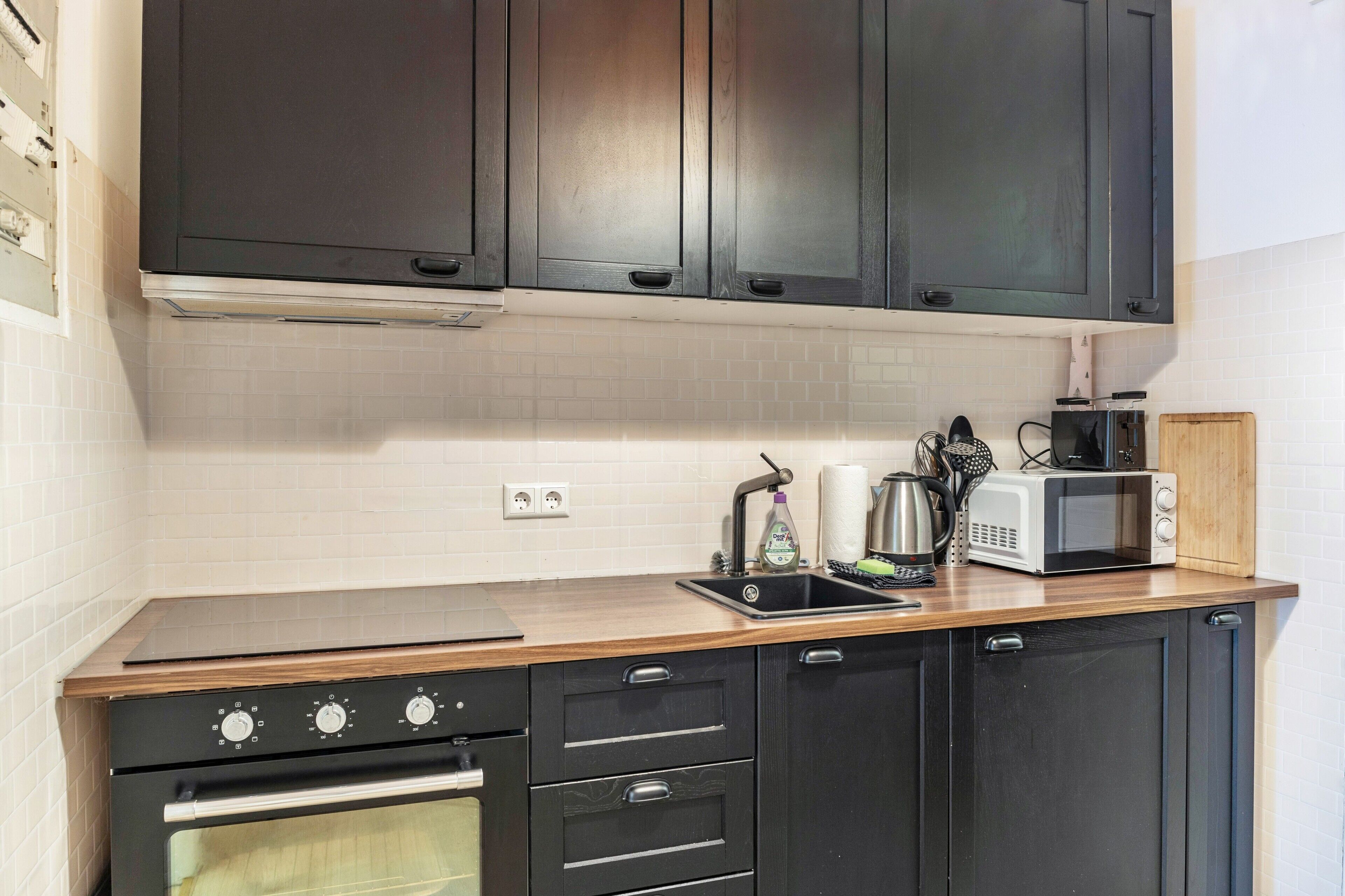 A kitchen with dark cabinets, a microwave, a kettle, and a wooden countertop.
