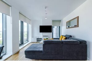Ein modernes Wohnzimmer mit einem grauen Sofa, einem Flachbildfernseher und großen Fenstern.