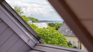 A view through a window with a roofline, a green bush, and a distant body of water. - Ty Annick – Les Glénans Room (Névez)