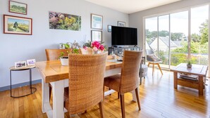 A dining area with wicker chairs, a wooden table, a TV, and framed pictures on the wall. - Ty Annick – Port Manech Room (Névez)