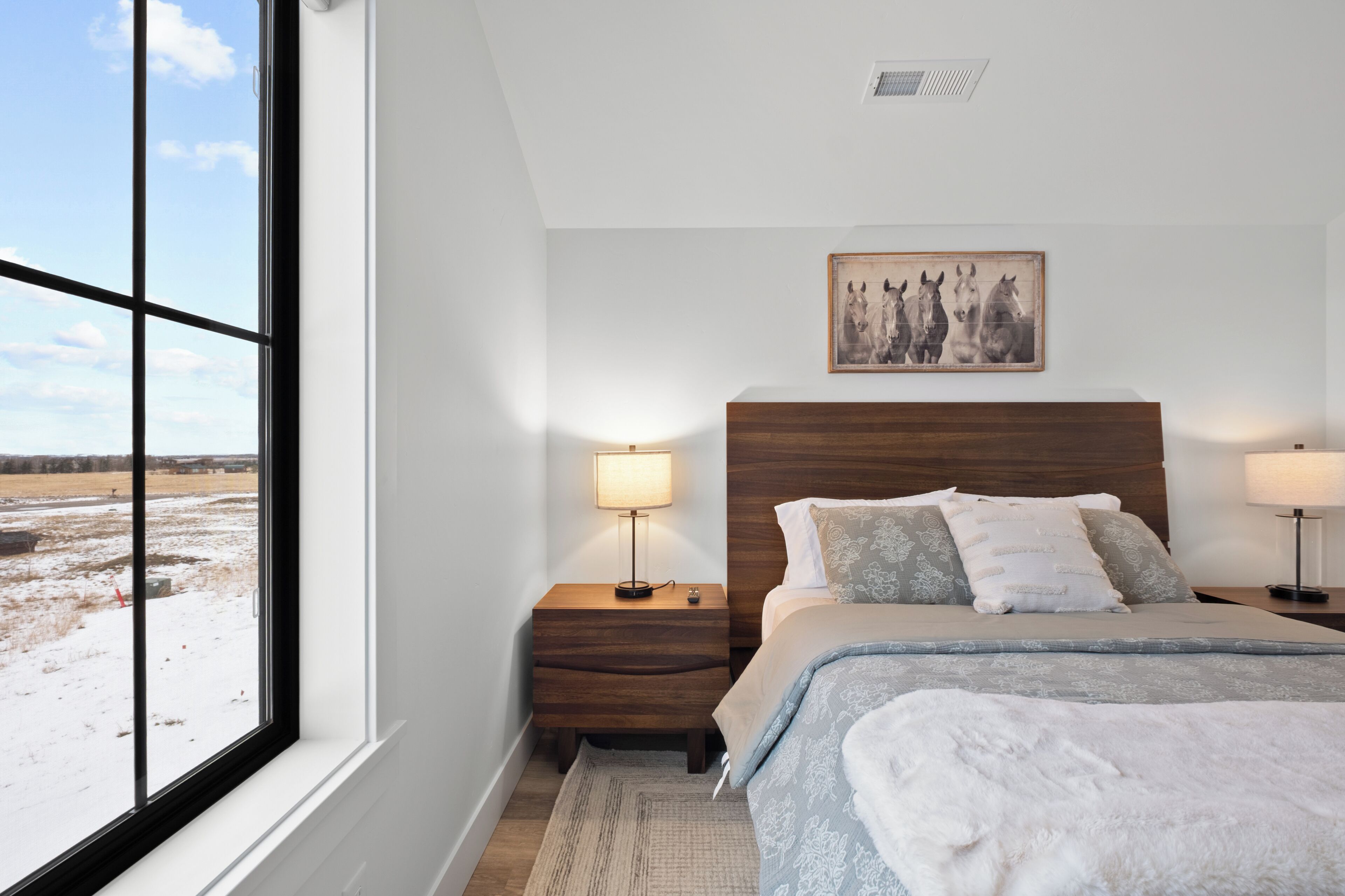 A bedroom with a large window, a wooden headboard, a bed with patterned bedding, two bedside tables with lamps, and a framed picture on the wall.