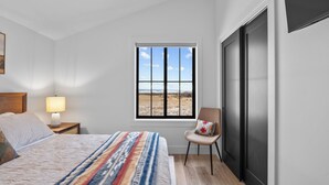 Un dormitorio con cama, mesita de noche con lámpara, silla, ventana con vista a un campo y puerta corrediza.