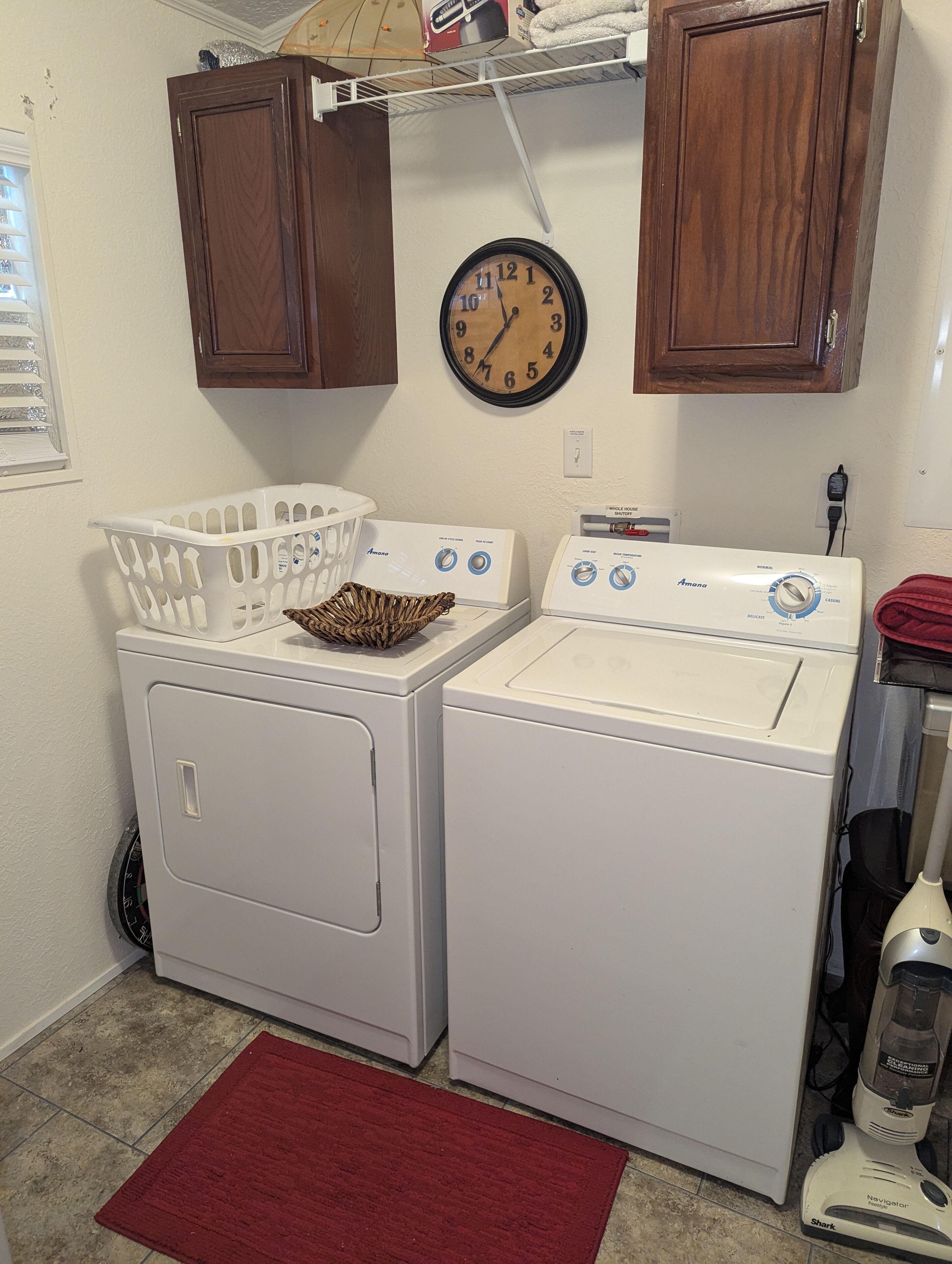 Une buanderie équipée de deux machines à laver à chargement frontal blanches, d’un sèche-linge blanc, d’une horloge, de meubles en bois et d’un tapis rouge.