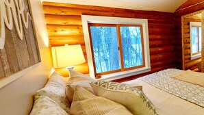 A cozy log cabin room with a bed, pillows, a bedside lamp, and a window with a view of trees. - Ely Lakeside Cabin: The Perfect Northwoods Retreat (Ely)