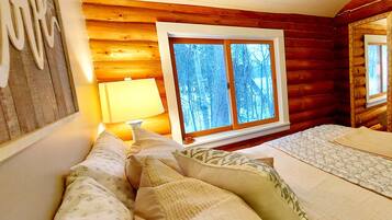 A cozy log cabin room with a bed, pillows, a bedside lamp, and a window with a view of trees.