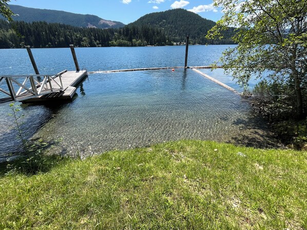 A clear lake with a wooden dock extending into the water, surrounded by grassy banks and trees. - Lakefront house with private dock (Lake Cowichan)