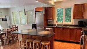 A modern kitchen with wooden cabinets, a central island with stools, a built-in oven, and a large window with a view of trees. - Lakefront house with private dock (Lake Cowichan)
