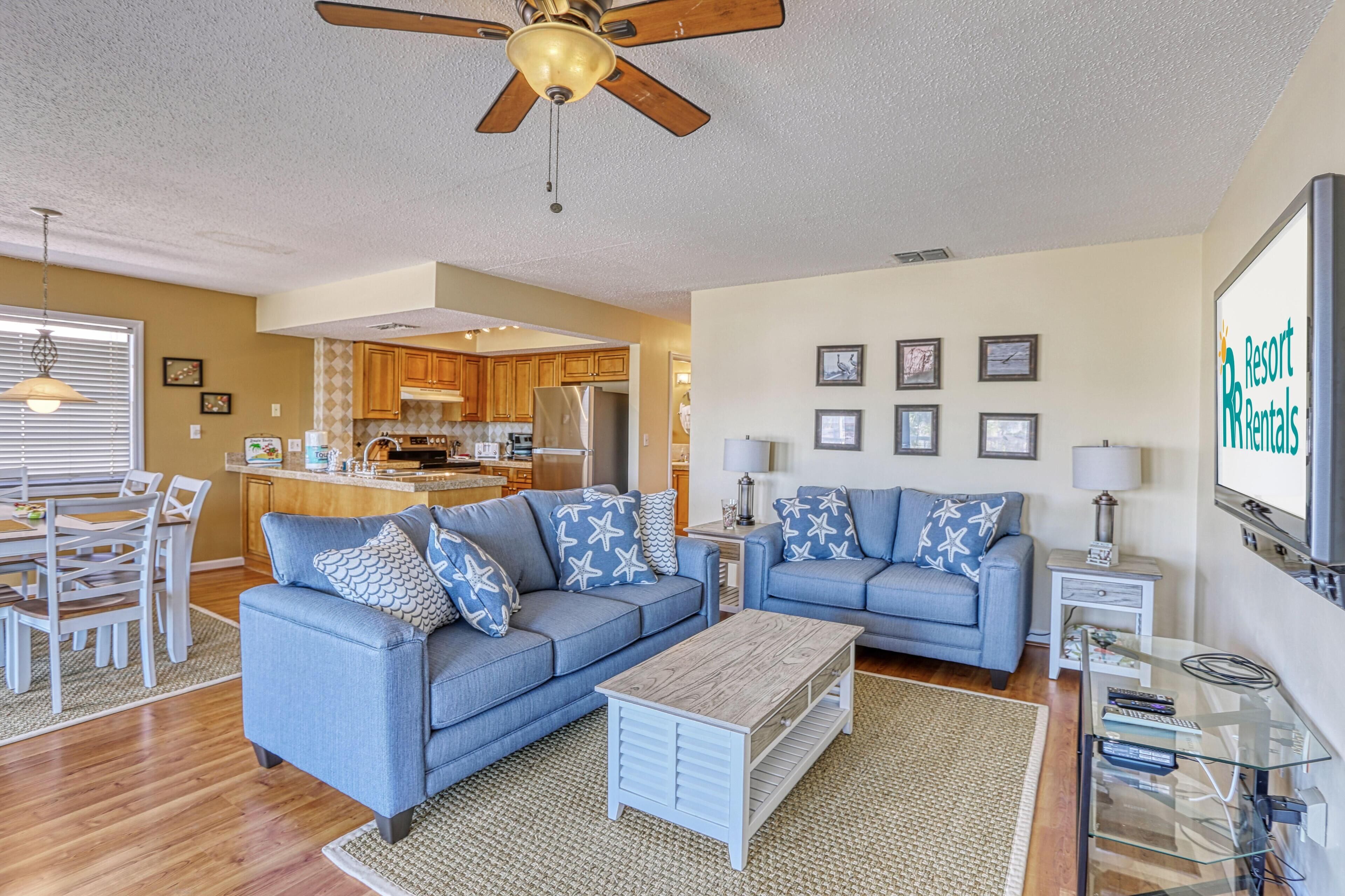 A spacious living room with two blue sofas, a wooden coffee table, and a ceiling fan. A kitchen with wooden cabinets and a dining area with a white table and chairs are visible in the background.