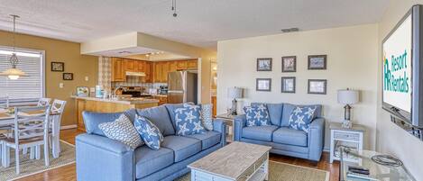 A spacious living room with two blue sofas, a wooden coffee table, and a ceiling fan. A kitchen with wooden cabinets and a dining area with a white table and chairs are visible in the background.