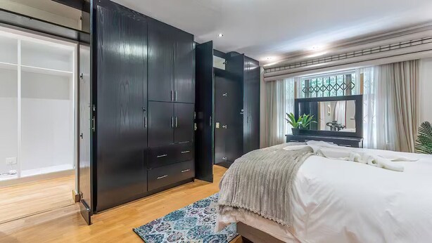A bedroom with a large wardrobe, a bed with a white comforter, a patterned rug, and a window with curtains. - Family Suite - Indoor Hot Tub, Heated Pool, Play Park & Table Mountain Views (Cape Town)