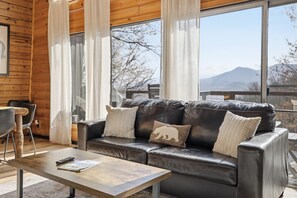 Un sofá de cuero con dos almohadones, una mesa de madera y vistas a montañas a través de grandes ventanales.