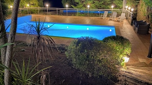 Une vue nocturne d’une piscine rectangulaire, bordée d’une terrasse carrelée et entourée d’une végétation luxuriante, avec des espaces de détente en plein air.