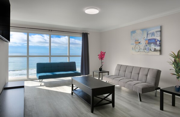 A modern living room with a large window overlooking the ocean, a blue sofa, a grey sofa, a coffee table, and a painting of a van and surfboard. - Monthly Rental in Brand New Boardwalk Condo (Myrtle Beach)