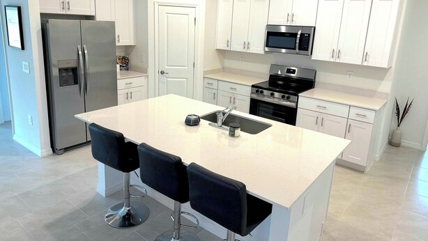 Una cocina moderna con armarios blancos, un refrigerador de acero inoxidable, un microondas, una estufa y un fregadero. Hay tres taburetes negros en la isla.