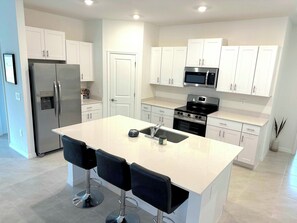 Una cocina moderna con armarios blancos, un refrigerador de acero inoxidable, un microondas, una estufa y un fregadero. Hay tres taburetes negros en la isla.