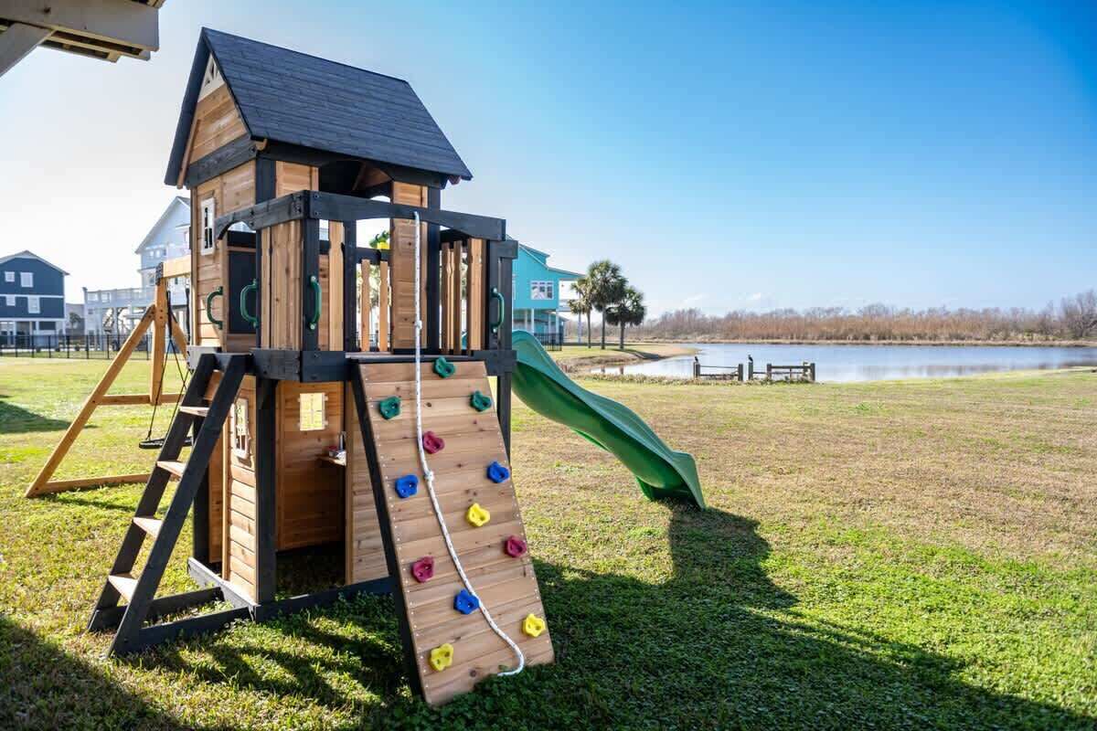Une structure de jeux en bois, dotée d’un toboggan et d’un mur d’escalade, installée sur une pelouse près d’une étendue d’eau.