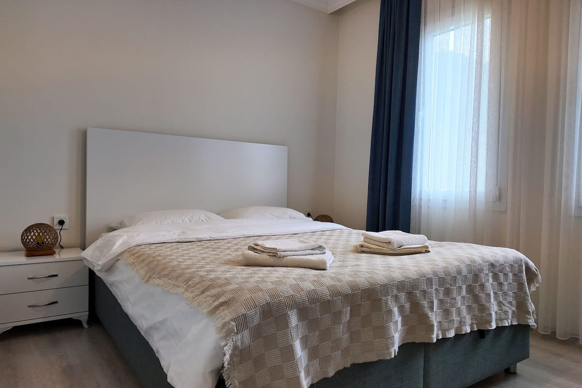 A neatly made bed with a patterned blanket, a bedside table with a lamp, and a window with sheer curtains.