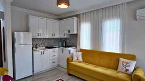 Eine kleine Küche mit weißen Schränken, einem gelben Sofa und einem Fenster mit transparenten Vorhängen.