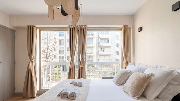 Ein Hotelzimmer mit einem großen Bett, beigen Vorhängen, Blick auf Gebäude und einer Pendelleuchte.