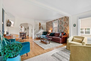 Een ruime woonkamer met een houten vloer, een blauwe fauteuil, een bruine leren bank en een beige fauteuil. Een trap leidt naar een verdieping.